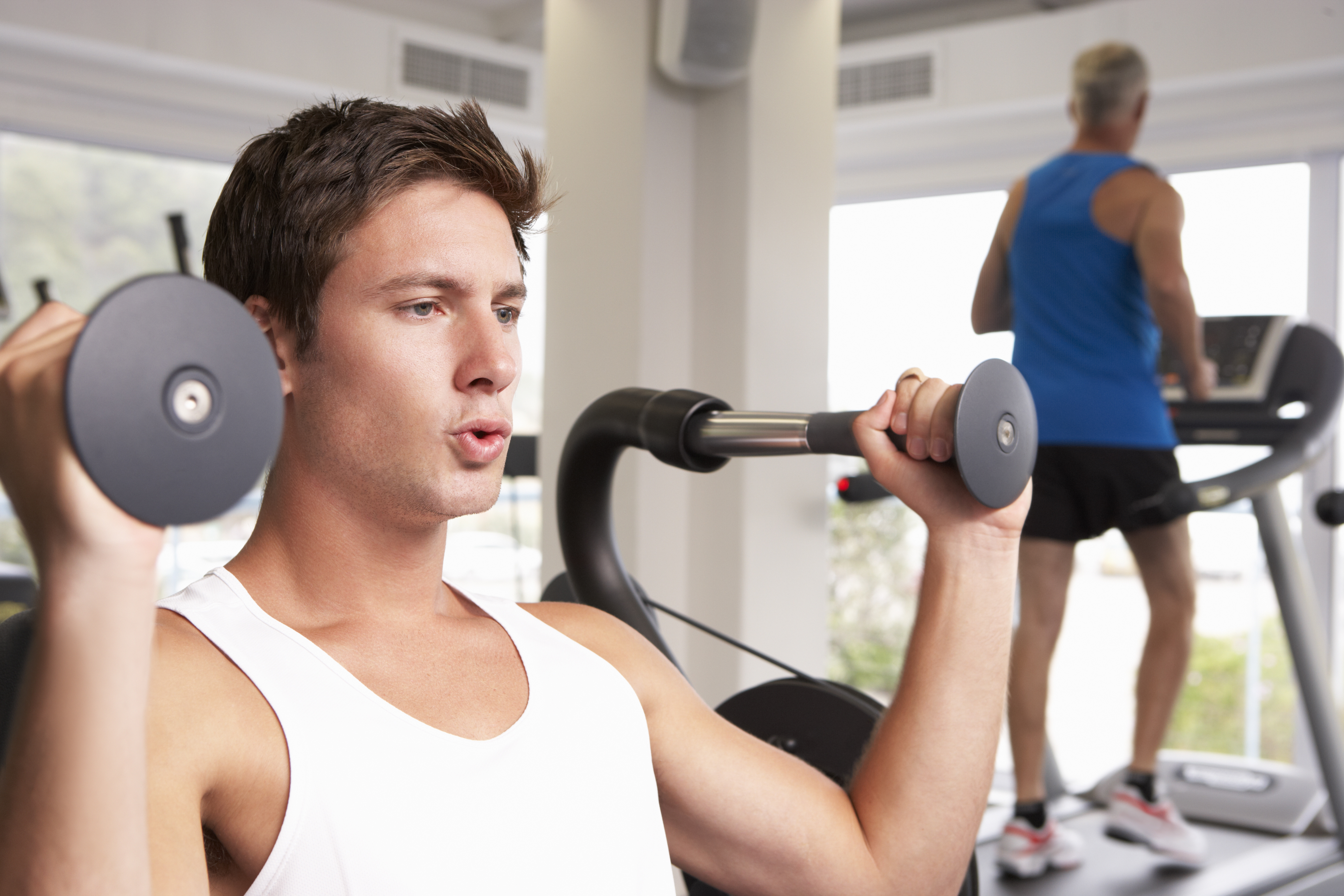 Man Using Weights Machine With Runner On Treadmill In Background